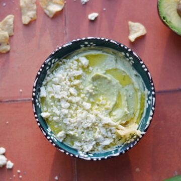Avocado feta dip in a blue bowl on a red background next to chips.