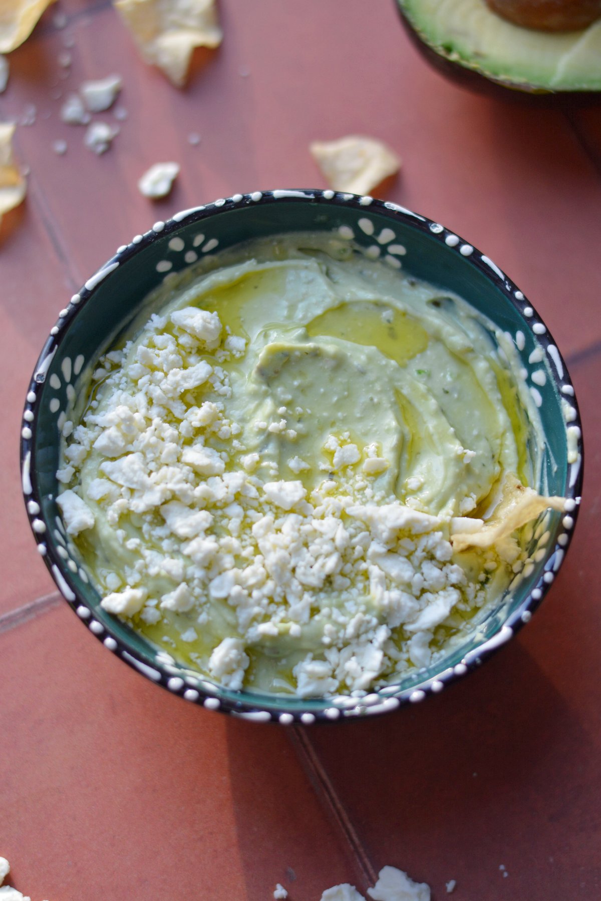 Avocado feta dip in a blue bowl with feta and oil on top on a red background next to chips.